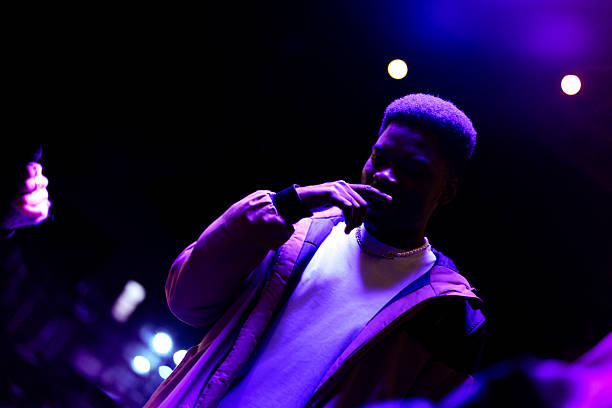 A focused young man stands in low light, surrounded by colorful stage lights at a lively urban music event. The atmosphere is energetic and dynamic, capturing the essence of nightlife.