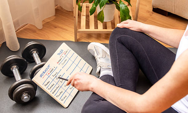 istock Woman sitting on a yoga mat is planning her weekly workout routine, using a clipboard and pencil, with dumbbells nearby, demonstrating her commitment to fitness and a healthy lifestyle 2211387565