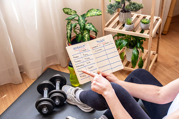 istock Woman sitting on a yoga mat pointing at her workout plan, ready to start exercising with dumbbells in her living room, surrounded by houseplants 2211387574