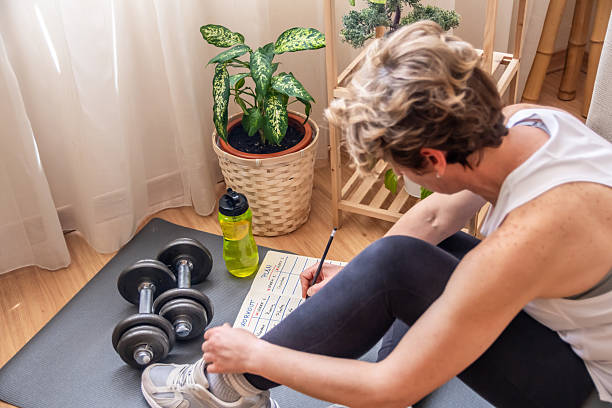istock Mature woman sitting on yoga mat, writing on workout plan, preparing for exercise with dumbbells at home, promoting healthy lifestyle and fitness routine 2211387577