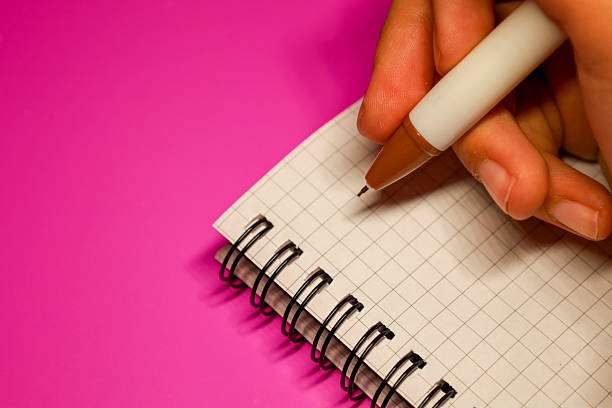 A human hand holds a white pen with a brown cap, ready to write in a wire-bound notebook. The notebook is open to a blank graph paper page.