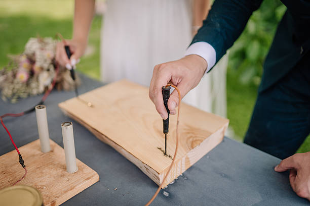 Close-up of two people woodburning on a wooden plank using pyrography tools, outdoors on a table. A bouquet and copy space are visible.