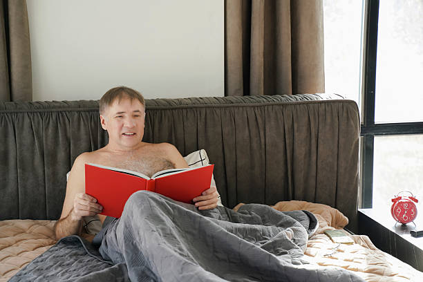 istock Man relaxed in bed on a calm morning, engaged in reading a red book while under a soft blanket 2211392247