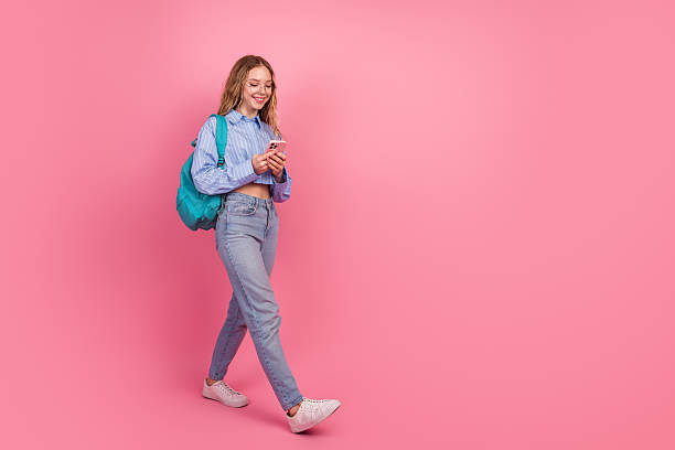 istock Young student walking with a backpack and phone against a pink background, showcasing casual style and trendy fashion 2211392880