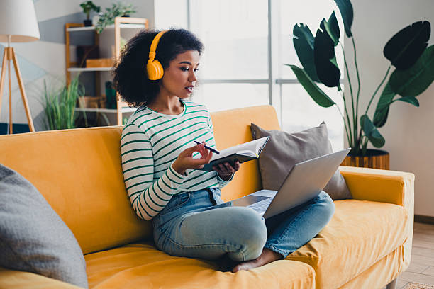 istock Young woman with headphones writing notes while on a laptop in a bright comfortable living room with plants decor 2211393455