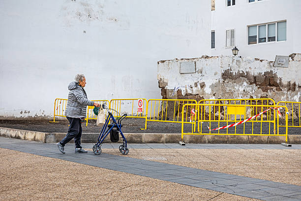 Arrecife, Lanzarote, Canary Islands, Spain - March 1st 2025:  An elderly woman walks steadily along a paved sidewalk, using her rollator for support. Dressed in a grey jacket and dark pants, she moves with quiet determination past a white wall with peeling paint, a sign of the passage of time. Yellow safety barriers surround a damaged section of the pavement, emphasizing the urban infrastructure and the precautions taken to ensure accessibility. The image captures a moment of resilience, independence, and the everyday challenges of navigating city streets.