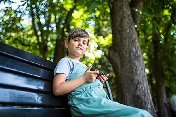 Girl sitting on a park bench, using a smartphone to browse social media and enjoying the warmth of a sunny summer day