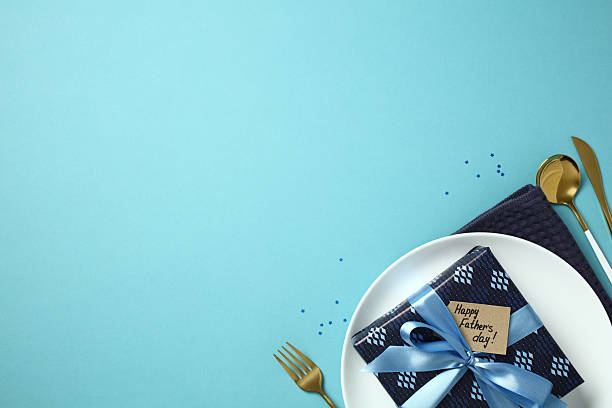 Father's Day celebration concept. Flat lay table setting with gift box on plate, golden cutlery, napkin, and greeting tag on a blue background.