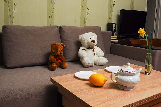 Two teddy bears sit on a couch next to a table set for tea, including a lemon and a daffodil.