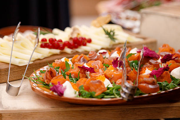 Close-up of a beautifully presented plate of smoked salmon appetizers with cream cheese and fresh herbs, alongside a cheese platter garnished with red currants and rosemary, on a wooden buffet table. The image highlights gourmet finger food, ideal for events, parties, and catering. It showcases the culinary delights and elegant food presentation often found in European cuisine and gatherings.