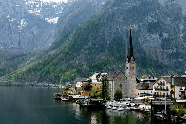 Scenic view of Hallstatt village with historic church traditional buildings alongside lake surrounded by majestic Alps mountains in Austria. Tourist boat docked at wooden pier.