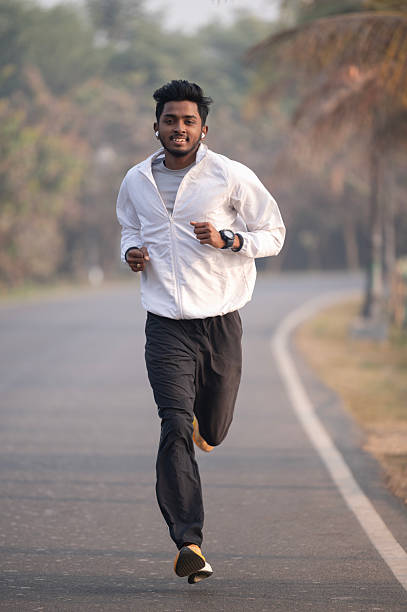 A young man jogs confidently along a peaceful road surrounded by nature. Dressed in a white sports jacket, black track pants, and orange running shoes, he represents dedication to fitness and a healthy lifestyle in an outdoor setting.