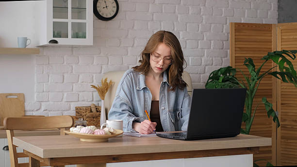 Seated at a kitchen table, a woman in glasses writes in a notebook while using a laptop. This photo captures the essence of modern remote work and the importance of a conducive workspace at home.