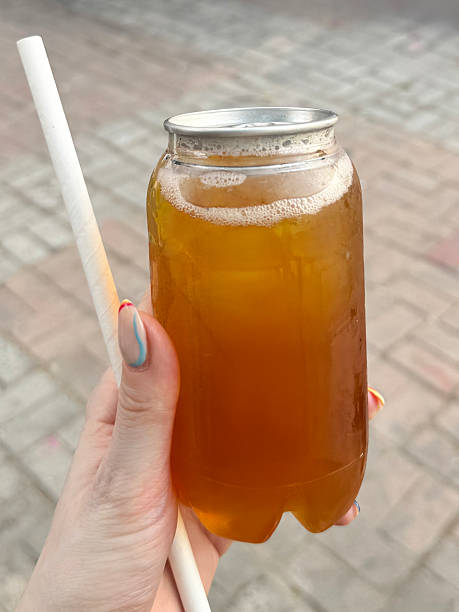 Close-up image of unrecognisable woman holding unopened, transparent plastic, single-use, disposable, ring pull can of iced tea and paper drinking straw, displaying nail art, brick paved yard background, focus on foreground