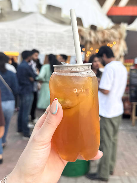 istock Close-up image of unrecognisable woman holding opened transparent plastic, single-use, disposable, ring pull can of iced tea, paper drinking straw, displaying nail art, crowds at outdoor food market setting, focus on foreground 2211421413