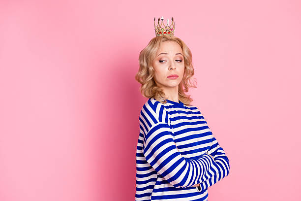 istock Confident Young Woman Wearing a Crown Displaying Attitude in a Striped Pullover in Front of a Pink Background 2211423126