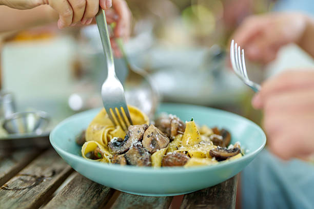 A close-up view of creamy mushroom pasta being twirled with a fork. The dish is served in a turquoise bowl atop a rustic wooden table, shared between two people.