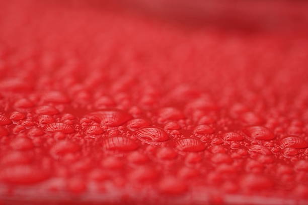 Close-up macro view of small water droplets scattered across a red surface, forming an organic and irregular texture. The shallow depth of field highlights the subtle variations in droplet size and pattern. The image conveys freshness and a vibrant, moist atmosphere through its strong color and detailed surface structure.