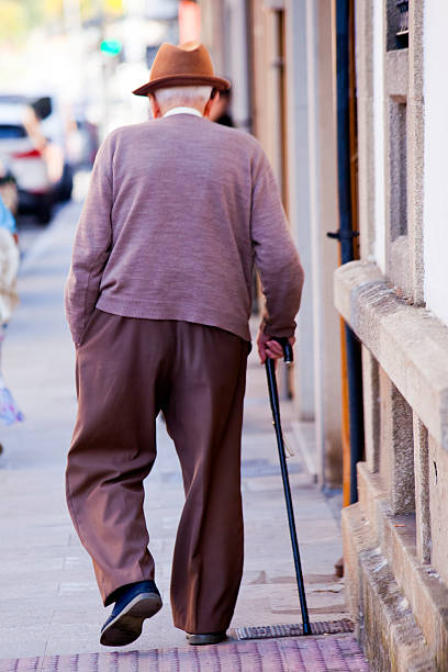 Disabled senior man wearing hat, walking in a hurry with walking cane along street sidewalk .  Galicia, Spain.