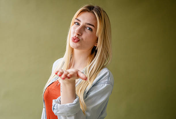 A young blonde woman smiles playfully while blowing a kiss in a stylish studio. Her cheerful demeanor and fashion sense highlight her individuality and confidence.