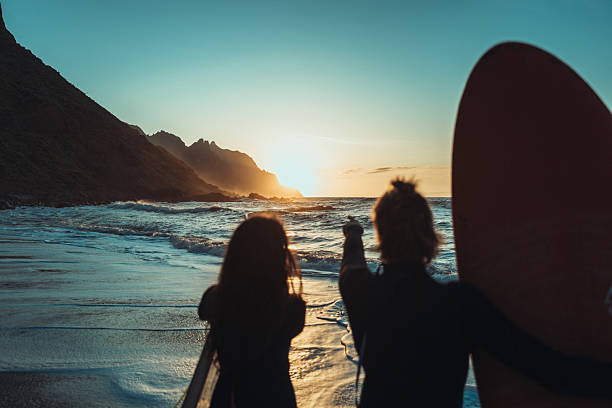 Two surfers walk toward the ocean at sunset on a black sand beach in Taganana, Tenerife, with dramatic cliffs and waves in the background.