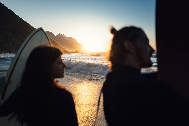 Two surfers walk toward the ocean at sunset on a black sand beach in Taganana, Tenerife, with dramatic cliffs and waves in the background.