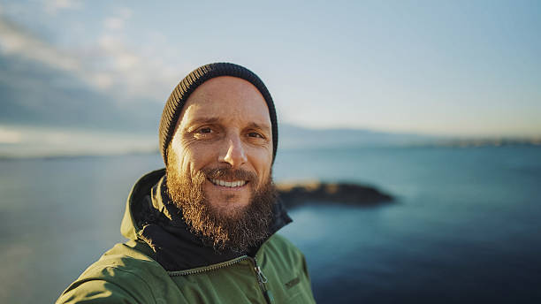 Man smiling and taking a selfie outdoors with a view of the ocean and distant mountains, wearing a green jacket and a black beanie.