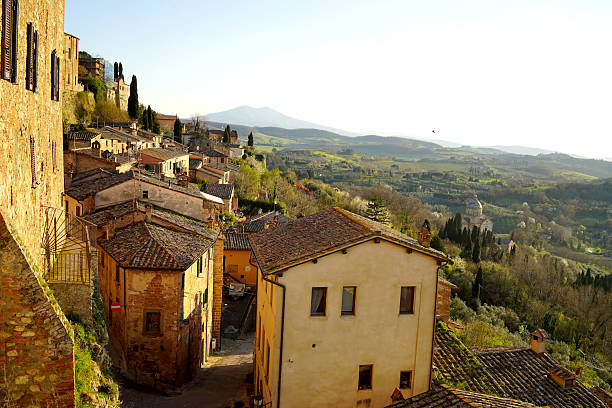 A stunning landscape featuring an expansive, cloudy blue sky dominating the scene above the rolling hills of Val D' Orcia, Tuscany. Soft, fluffy clouds cast dynamic shadows over the lush green fields below, creating a beautiful interplay of light and shadow. The serene countryside showcases the region's iconic undulating terrain, dotted with patches of cypress trees, villas aand vibrant greenery, evoking a sense of tranquility and natural beauty.