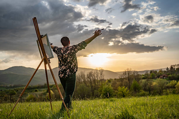 A man immerses himself in painting outdoors, capturing the vivid sunset over a lush landscape with mountains.