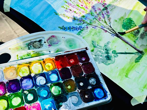 A close up an unrecognisable person dipping their yellow paintbrush into their watercolour paint pallet and art set which also includes colour pencils. The person is enjoying the beauty in nature and painting outside in the Lake District during springtime.