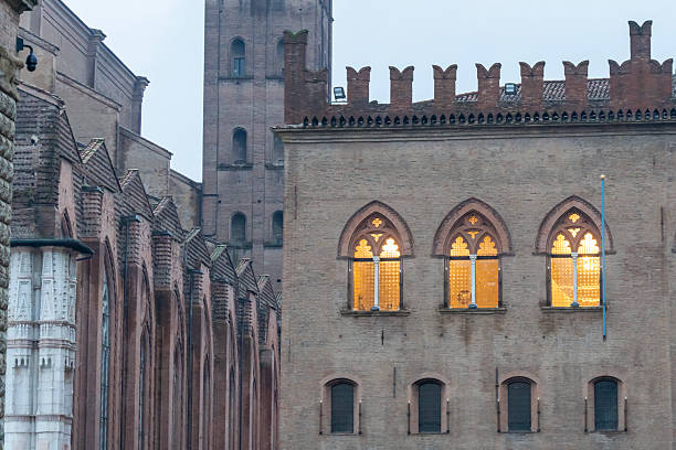 Historic brick buildings with pointed Gothic-style windows glow warmly from within during the evening. The scene captures the intricate architectural details and historic atmosphere of a Bologna, Italy