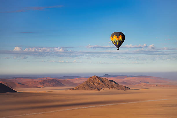 A vibrant hot air balloon ascends into the pale dawn sky over Namibia's vast desert plains. Below, a rocky hill rises from the sand, its rugged surface catching the soft glow of sunrise. The empty expanse and gentle color gradients offer a sense of freedom and wonder from above.