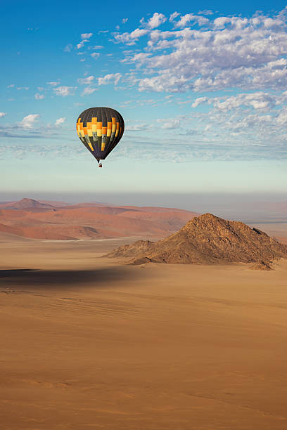 A brightly patterned hot air balloon floats above a desert plateau and rounded hills in Namibia, suspended in an open blue sky. The crisp atmosphere and wide-open space highlight the scale of the landscape below and the tranquility of ballooning far from civilization.
