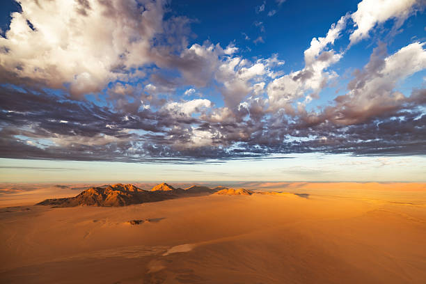 Golden desert stretches endlessly under a bright blue sky filled with soft, swirling clouds. Mountains rise in the distance, creating a serene and expansive scene in the open air.