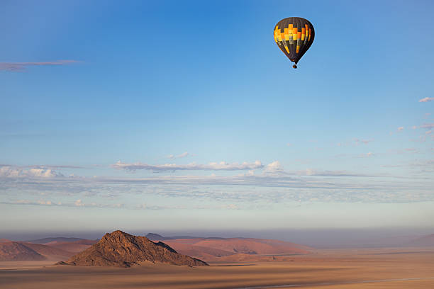 A single hot air balloon floats peacefully above a solitary desert peak in Namibia, framed by soft pastel skies. The early morning light casts long shadows across the sand and paints the surrounding hills in muted purples and oranges. The scene evokes isolation, stillness, and the quiet grandeur of flight over untouched terrain.