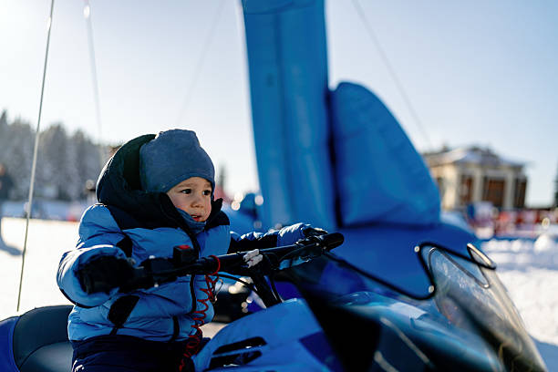 A young child dressed in warm winter clothing sits on a snowmobile in a scenic snowy setting. The child appears engrossed in the adventure, with a blue sky and snowy background completing the outdoor experience, conveying enjoyment and exploration during a winter holiday.