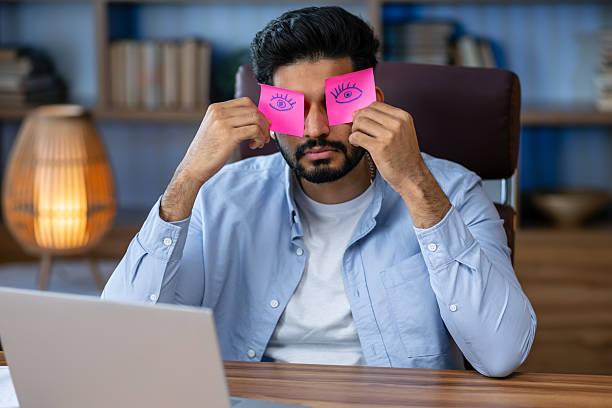 istock Exhausted tired businessman with painted eyes on stickers, adhesive notes on face sleeping at workplace, sitting at desk with laptop, unproductive lazy young male dozing, working on difficult project 2211508502