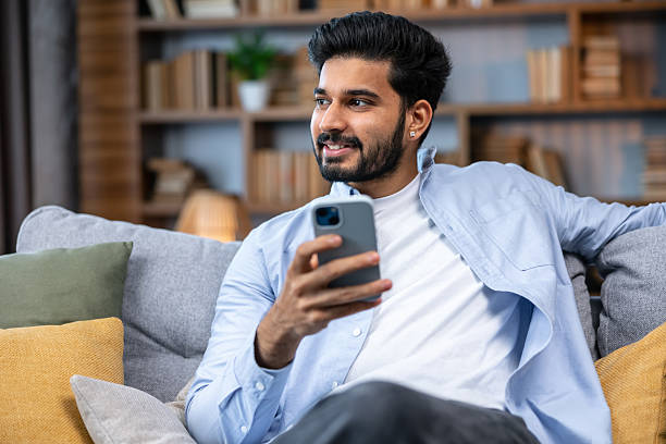 istock Attractive smiling young man relaxing on a couch at home, using mobile phone 2211508526