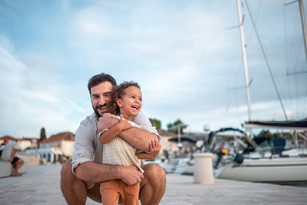 A father in casual attire embraces his joyful mixed race son at a small Sutivan harbour. The father smiles down at his son, capturing a moment of connection by the marina's serene setting, surrounded by boats and clear skies.