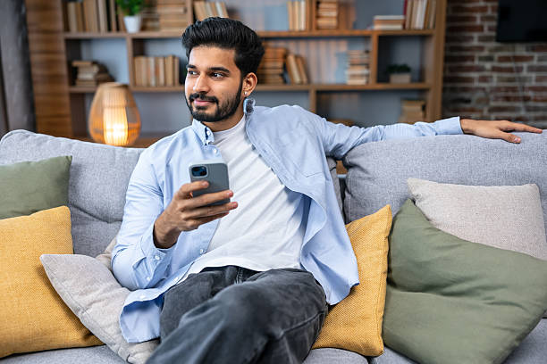 istock young man holding using cell phone sitting on sofa couch in living room. Cool guy browsing internet, surfing web 2211510224