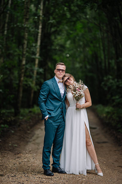 istock Bride and groom stand close together on a forest path, wearing sunglasses and smiling lovingly, with the bride holding a wildflower bouquet. 2211547049