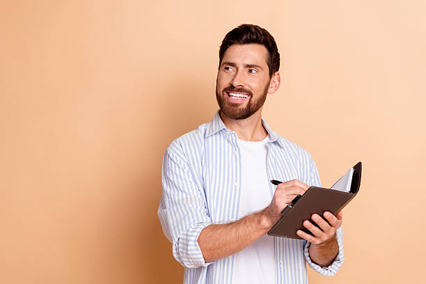 istock Young man smiling while writing in a notebook on a beige background wearing a blue striped shirt with a trendy appearance 2211548691