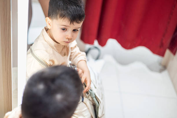 kid in fitting, dressing room trying on clothes. boy with stack of wears on hands sitting on chair. cute adorable child making funny faces in mirror.smiling baby in shopping center, mall.