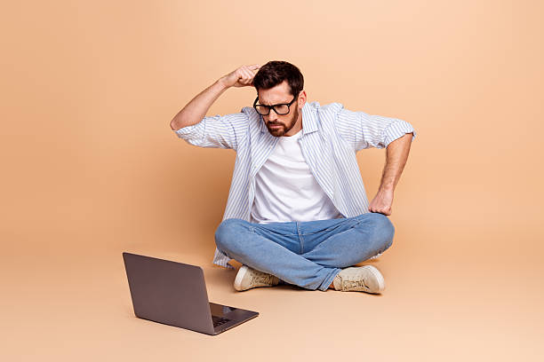 istock Young man sitting cross-legged on beige background while thinking and looking at laptop 2211549594
