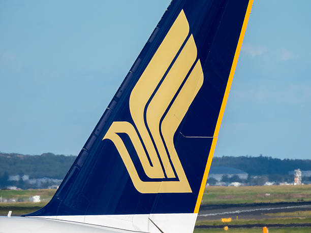 The vertical stabiliser of a Singapore Airlines Airbus A380-841 plane, registration 9V-SKY, taxiing for departure from Sydney Kingsford Smith Airport as flight SQ222 to Singapore. In the background is the main runway and in the far background are fuel storage tanks on the far side of Botany Bay. This image was taken from New Link Road, Mascot on a hot and cloudy afternoon on 21 April 2025.