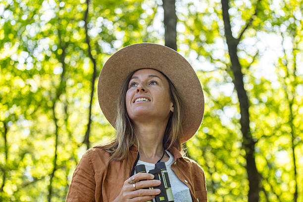 Woman in a sunlit forest wearing a hat, holding binoculars, and gazing upward. Curious explorer enjoying birdwatching in woodland. Outdoor adventure and nature discovery.