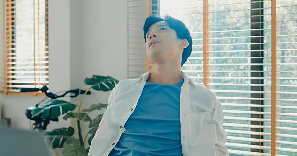 Closeup of young Asian man taking a break to stretch while working from home, emphasizing the importance of ergonomics and relaxation. Remote work and finance concept.