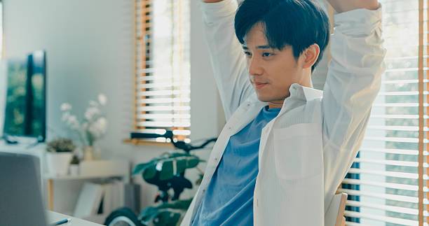 Young Asian man taking a break to stretch while working from home, emphasizing the importance of ergonomics and relaxation. Remote work and finance concept.