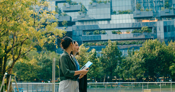 istock Two young Asian businesswomen discussing work while holds digital tablet and carries documents while walking outside office building. Sustainable business outdoor. 2211564635