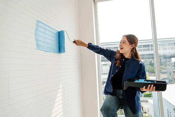 istock Young woman painting an interior wall light blue using a roller, smiling during a DIY home renovation. Natural daylight from a large window adds a bright, cheerful vibe to the project scene. 2211564972